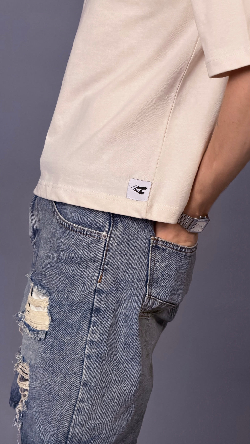 Person wearing a beige shirt with a logo and ripped blue jeans on a gray background