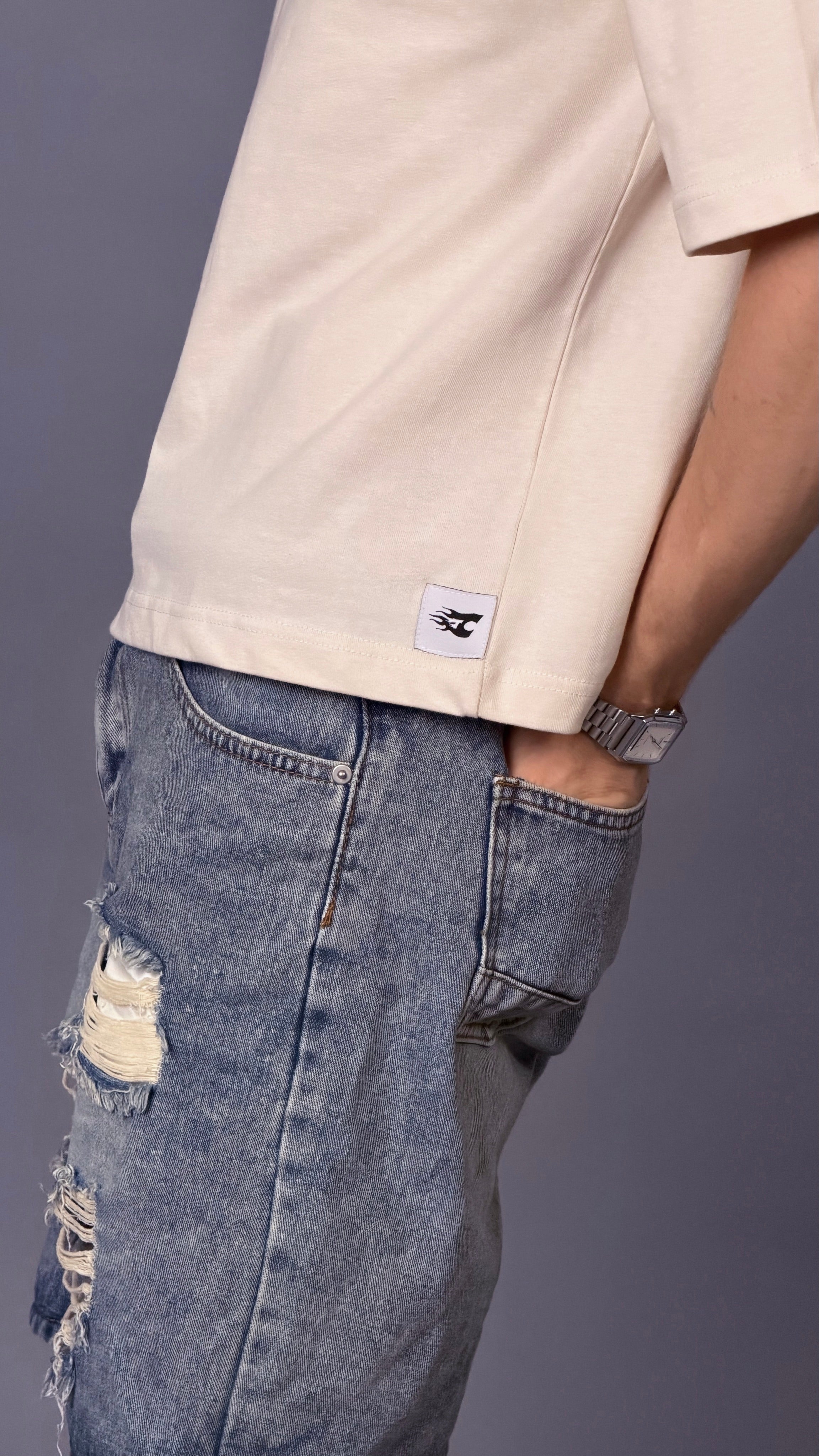 Person wearing a beige shirt with a logo and ripped blue jeans on a gray background
