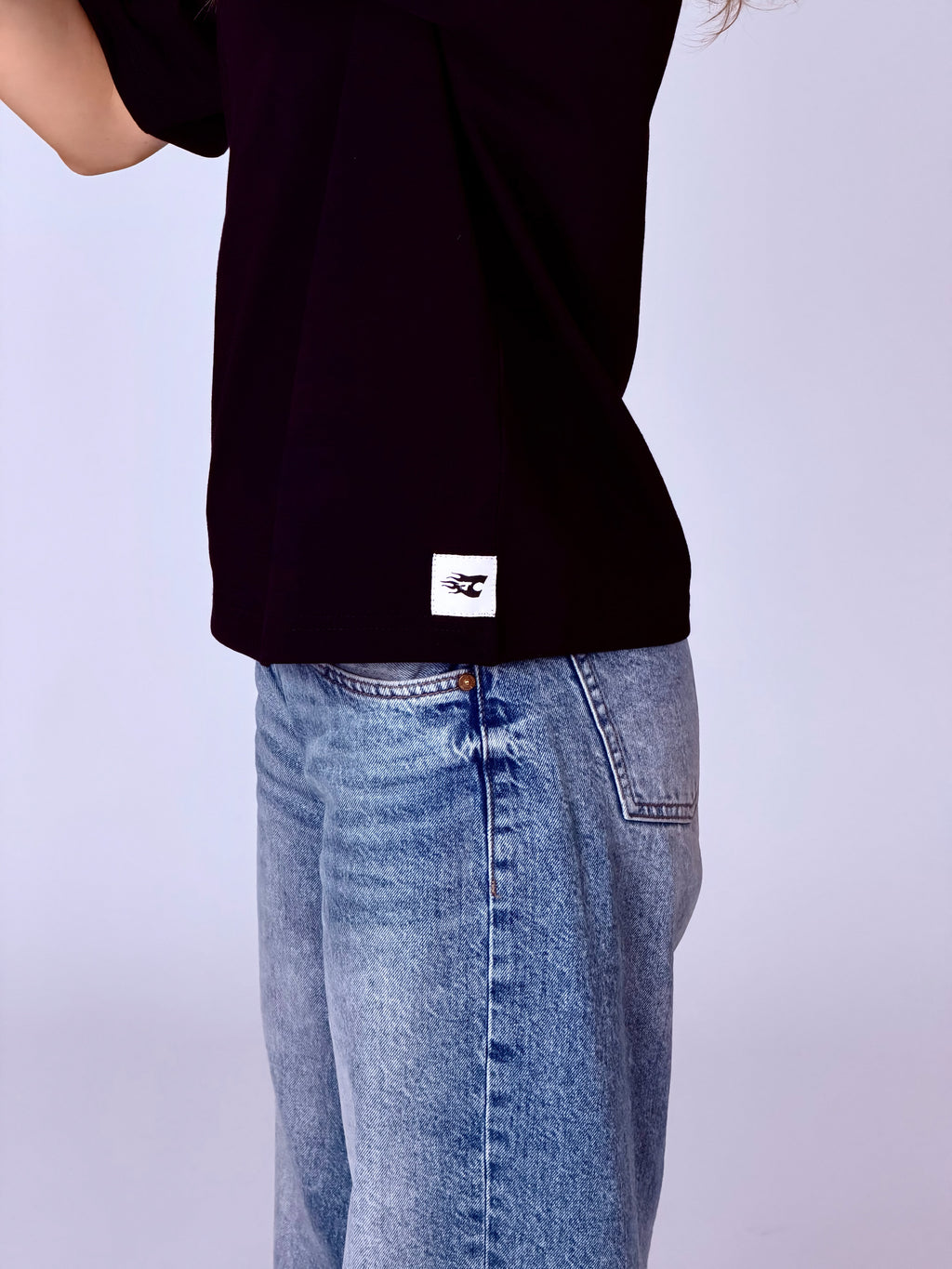 Person wearing a black t-shirt with a white logo and blue jeans on a light gray background