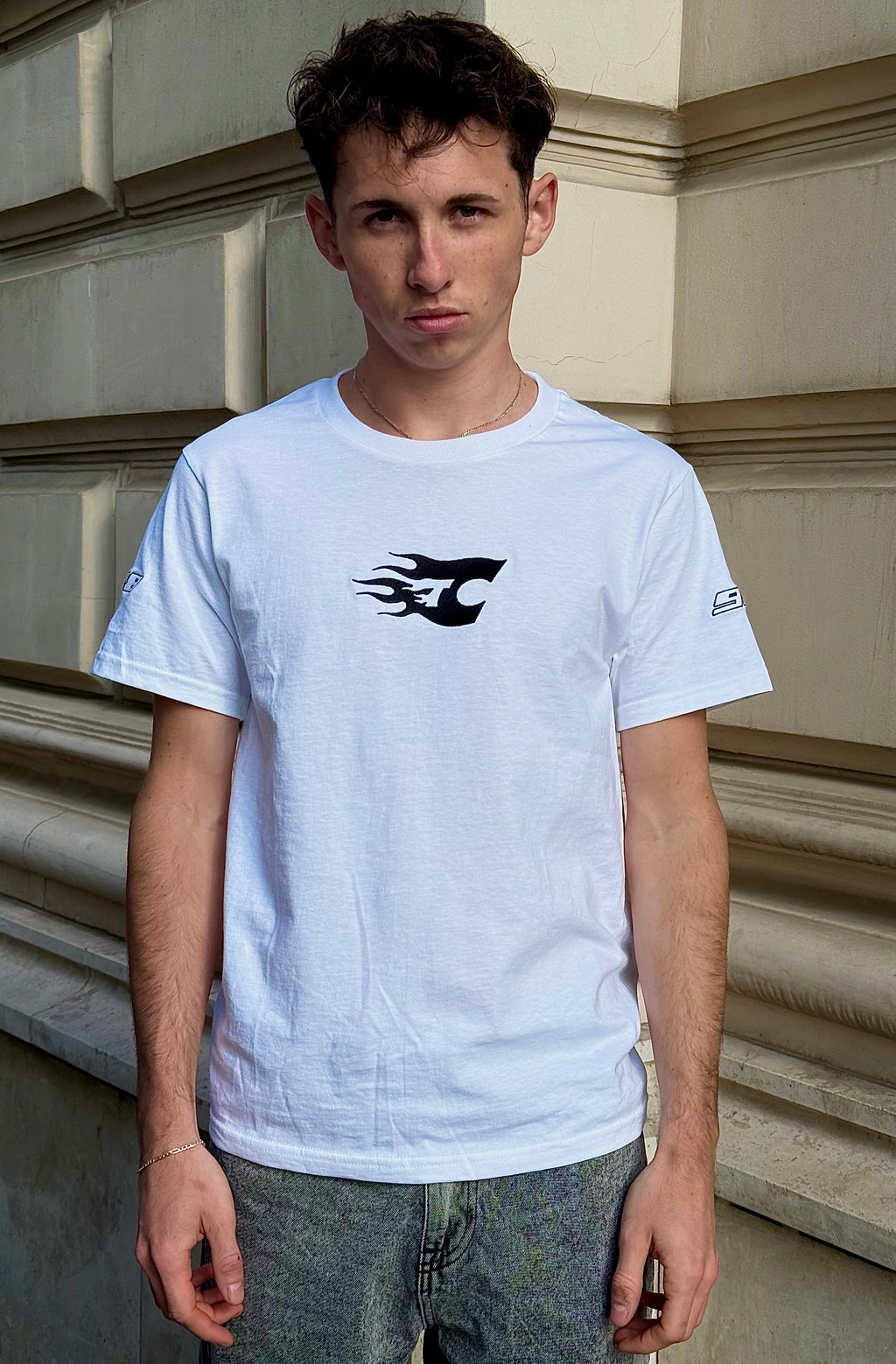 Person wearing a white t-shirt with a black logo against a beige wall.