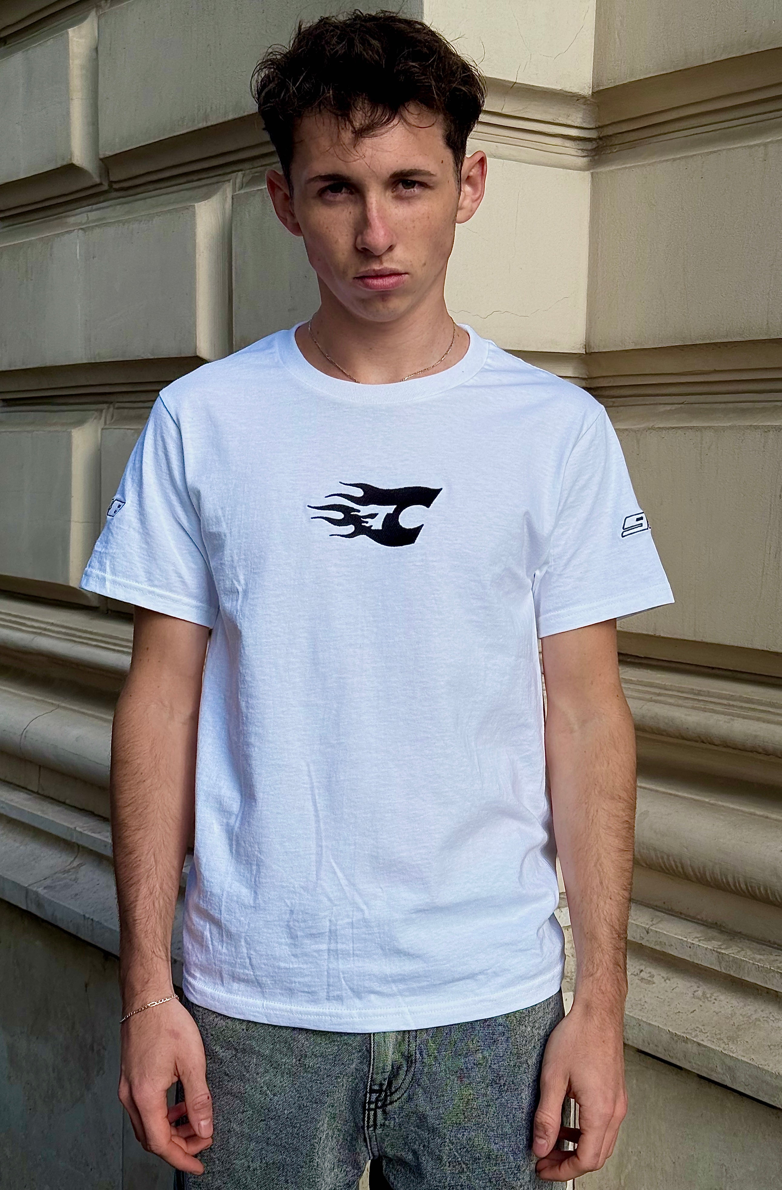 Person wearing a white t-shirt with a black logo against a beige wall.