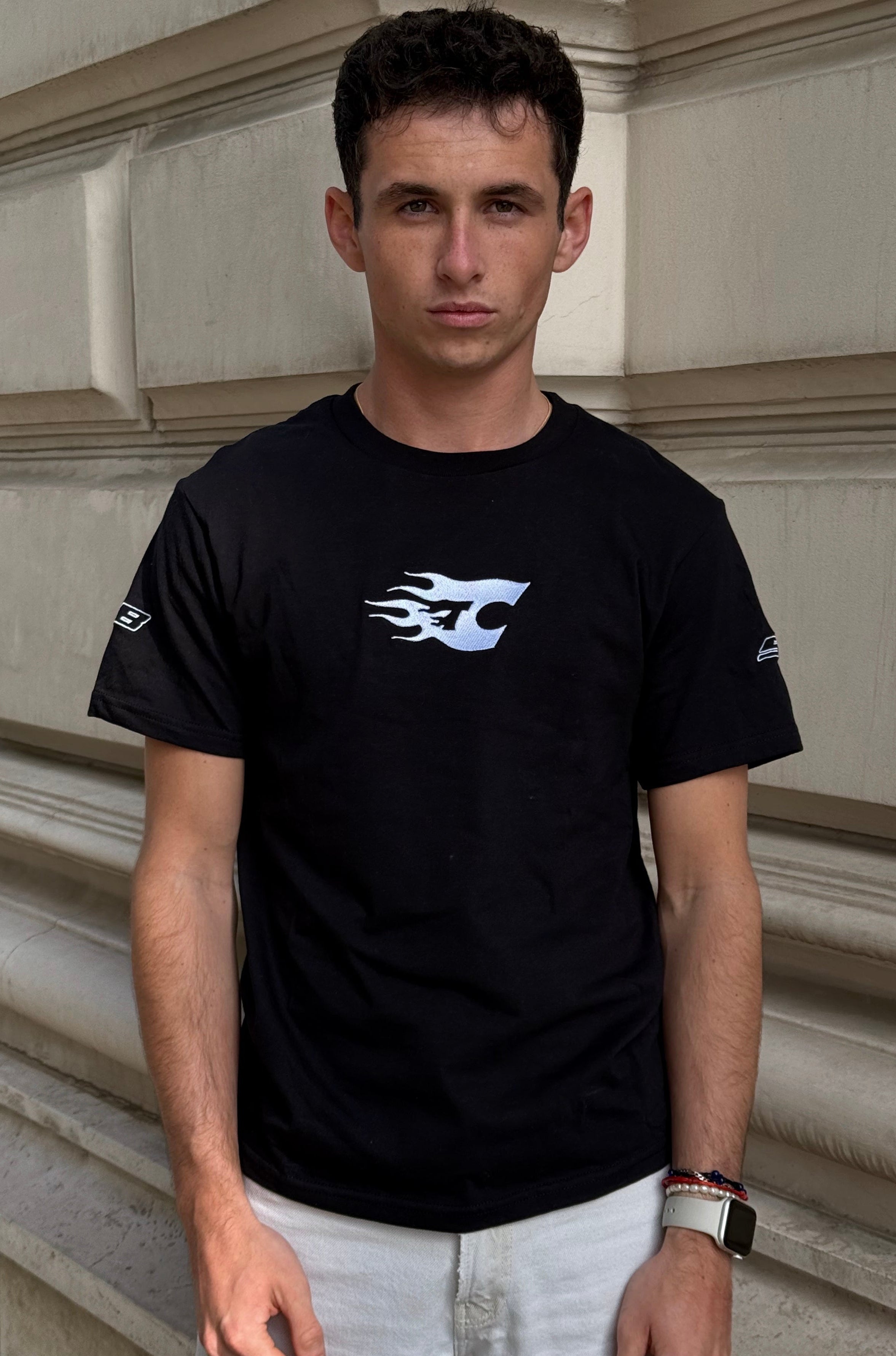 Man wearing a black t-shirt with a logo standing against a stone wall.