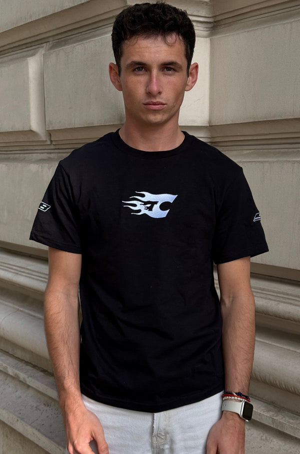 Man wearing a black t-shirt with a logo standing against a stone wall.