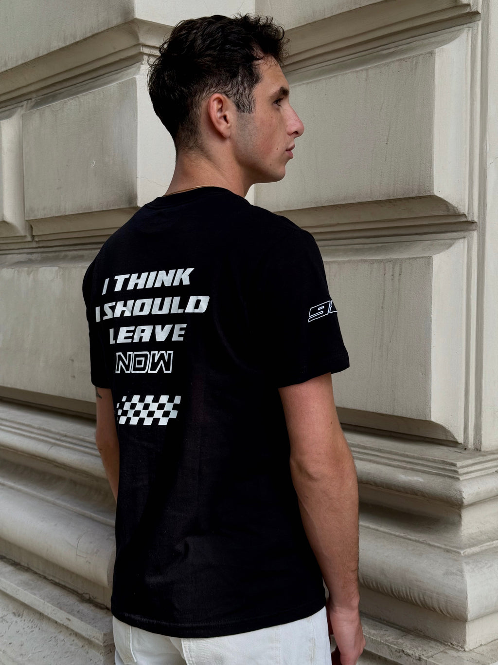 Man wearing a black t-shirt with text on the back, standing against a stone wall.