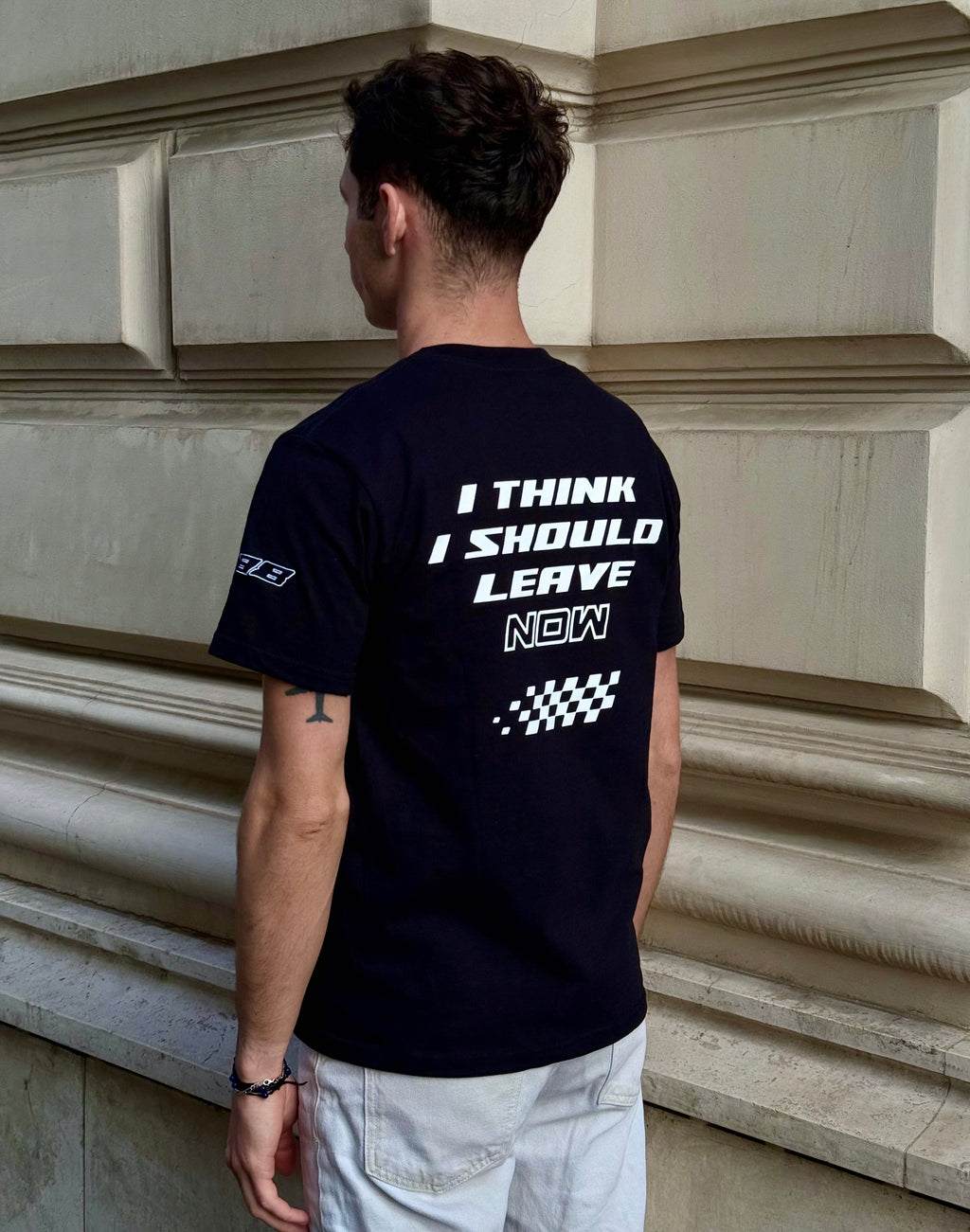Person wearing a black t-shirt with text on the back, standing against a stone wall.