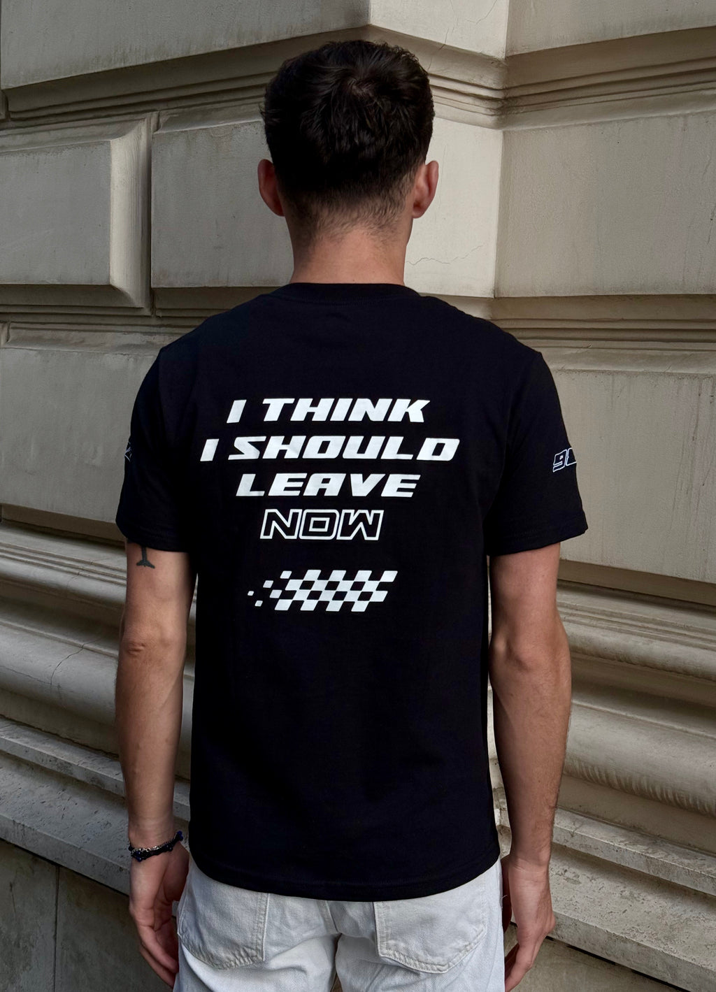Person wearing a black t-shirt with text on the back, standing against a building wall.