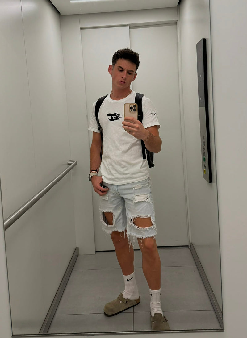 Person taking a mirror selfie in an elevator wearing a white t-shirt, ripped shorts, and a backpack.